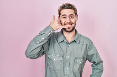 Hispanic man with beard standing over pink background smiling doing phone gesture with hand and fingers like talking on the telephone. communicating concepts. 