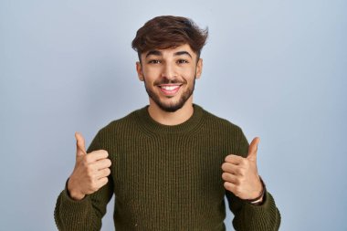 Arab man with beard standing over blue background success sign doing positive gesture with hand, thumbs up smiling and happy. cheerful expression and winner gesture. 