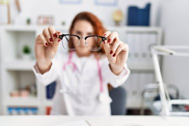 Hastanede elinde gözlük tutan doktor üniforması giyen genç kızıl saçlı kadın.
