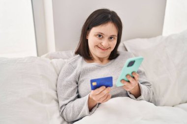 Yatak odasında yatakta oturan, akıllı telefon ve kredi kartı kullanan, Down sendromlu genç bir kadın.