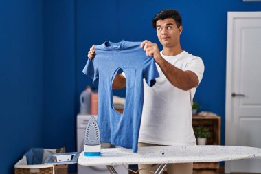 Hispanic man ironing holding burned iron shirt at laundry room smiling looking to the side and staring away thinking. 