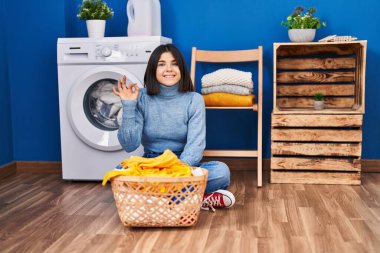 Young hispanic woman at laundry room doing ok sign with fingers, smiling friendly gesturing excellent symbol 