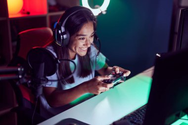 Young beautiful hispanic woman streamer playing video game using joystick at gaming room