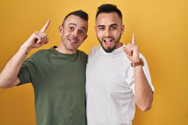 Homosexual couple standing over yellow background pointing finger up with successful idea. exited and happy. number one. 