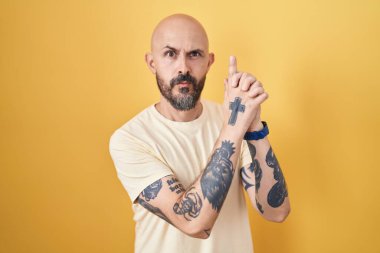 Sarı arka planda dikilen İspanyol dövmeli bir adam. Elinde sembolik bir silahla el hareketleriyle cinayet silahlarıyla oynuyor. Kızgın bir yüz. 