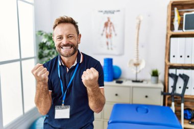 Middle age physiotherapist man working at pain recovery clinic very happy and excited doing winner gesture with arms raised, smiling and screaming for success. celebration concept. 