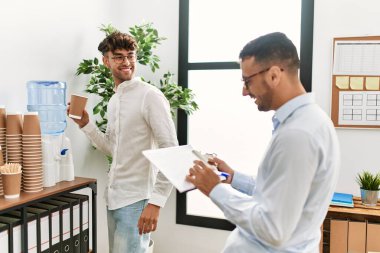 Two hispanic men business workers drinking water and writing on clipboard working at office