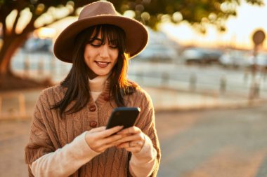 Kışlık şapkalı esmer kadın gün batımında şehrin dışında akıllı telefon kullanarak gülümsüyor.