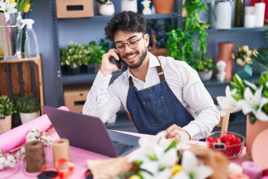 Young hispanic man florist using laptop talking on smartphone at florist shop