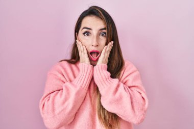 Pembe arka planda duran genç İspanyol kadın. Korkmuş ve şok olmuş, şaşırmış ve hayrete düşmüş. Yüzünde elleri olan. 