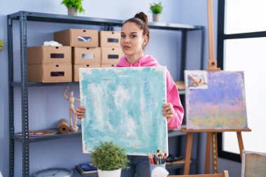 Young hispanic girl holding canvas relaxed with serious expression on face. simple and natural looking at the camera. 