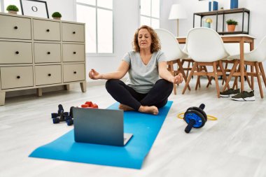 Orta yaşlı beyaz bir kadın evde laptopla yoga eğitimi veriyor.