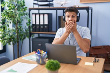 Young hispanic man working at the office wearing headphones shocked covering mouth with hands for mistake. secret concept. 
