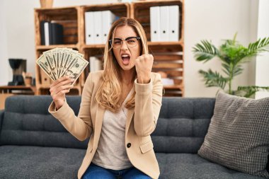 Young blonde therapist woman working at therapy office holding money annoyed and frustrated shouting with anger, yelling crazy with anger and hand raised 