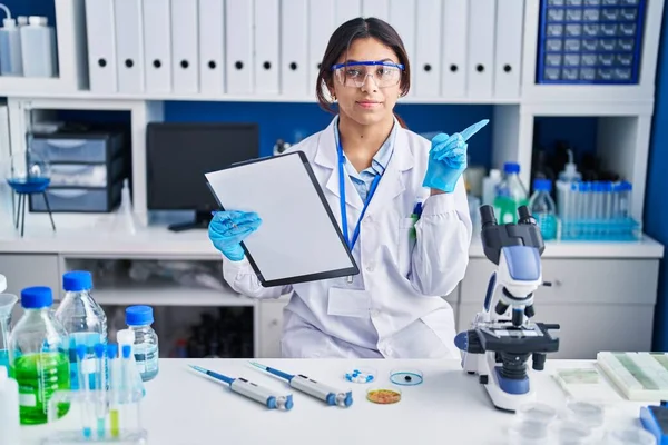 Hispanic young woman working at scientist laboratory pointing with hand finger to the side showing advertisement, serious and calm face