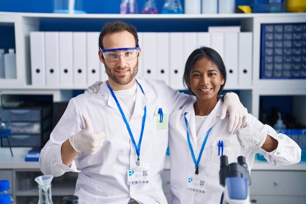Interracial couple working at scientist laboratory smiling happy and positive, thumb up doing excellent and approval sign