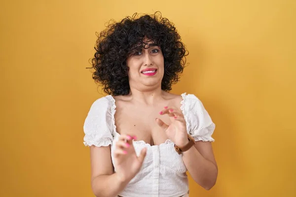 Young brunette woman with curly hair standing over yellow background disgusted expression, displeased and fearful doing disgust face because aversion reaction.