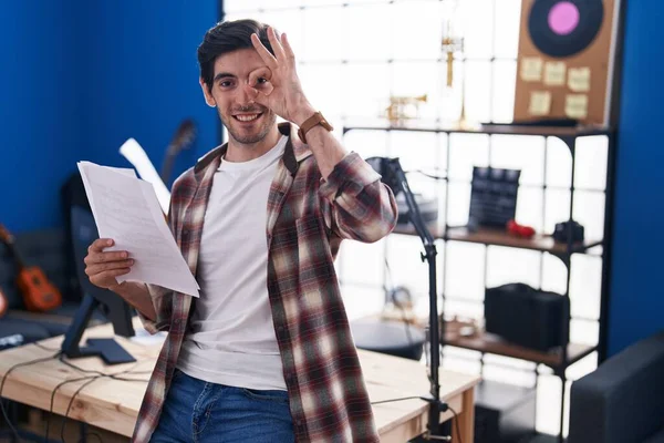 Young hispanic man reading music sheet at music studio smiling happy doing ok sign with hand on eye looking through fingers