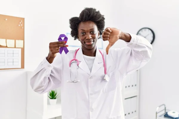 African doctor woman holding purple ribbon awareness at medical clinic with angry face, negative sign showing dislike with thumbs down, rejection concept