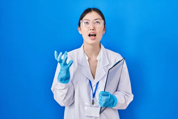 Chinese young woman working at scientist laboratory crazy and mad shouting and yelling with aggressive expression and arms raised. frustration concept.