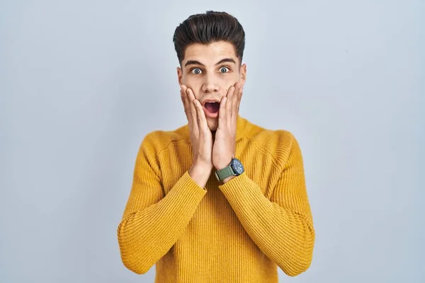 Young hispanic man standing over blue background afraid and shocked, surprise and amazed expression with hands on face