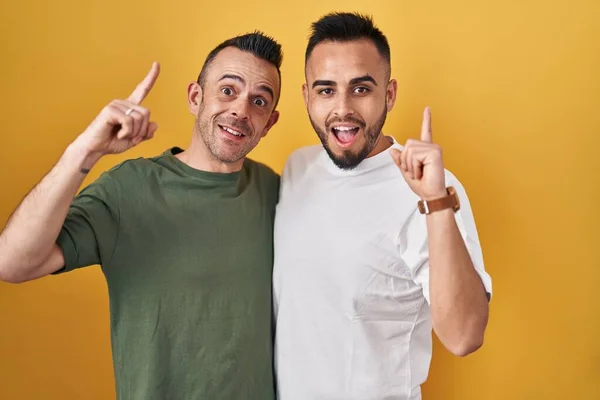 Homosexual couple standing over yellow background pointing finger up with successful idea. exited and happy. number one.