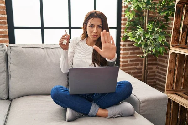Young brunette woman using laptop holding keys of new home with open hand doing stop sign with serious and confident expression, defense gesture