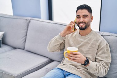 Genç Arap adam kahve içiyor ve evde akıllı telefondan konuşuyor.