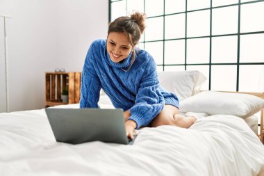 Young beautiful hispanic woman using laptop sitting on bed at bedroom