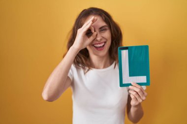 Beautiful brunette woman holding l sign for new driver smiling happy doing ok sign with hand on eye looking through fingers 