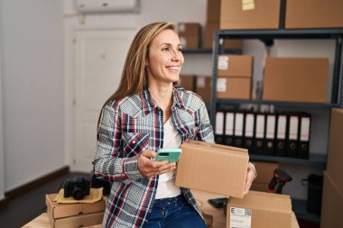 Genç sarışın kadın ecommerce iş işçisi ofiste akıllı telefon paketi kullanıyor.