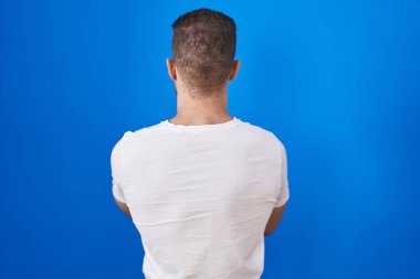 Young caucasian man standing over blue background standing backwards looking away with crossed arms 