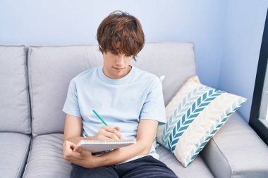 Young blond man writing on notebook sitting on sofa at home