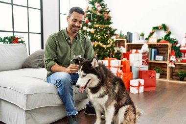 Genç İspanyol adam evdeki Noel ağacının yanında köpekle birlikte kanepede şarap içiyor.