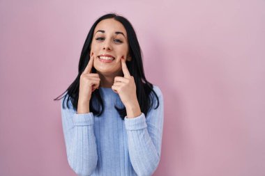 Hispanic woman standing over pink background smiling with open mouth, fingers pointing and forcing cheerful smile 