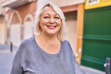 Orta yaşlı sarışın kadın sokakta kendinden emin bir şekilde gülümsüyor.