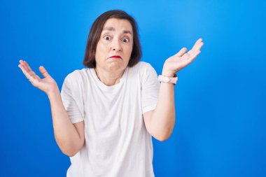 Orta yaşlı İspanyol kadın mavi arka planda duruyor. Kolları ve elleri havada, kafası karışmış bir ifade. şüphe kavramı. 