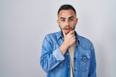 Young hispanic man standing over isolated background looking fascinated with disbelief, surprise and amazed expression with hands on chin 
