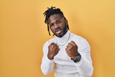 Sarı arka plan üzerine balıkçı yaka kazak giyen Afrikalı bir adam. Yumruk savunma hareketleriyle dövüşmeye hazır, kızgın ve üzgün yüzlü, sorundan korkan. 