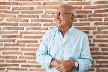 Senior man with grey hair standing over bricks wall looking away to side with smile on face, natural expression. laughing confident. 