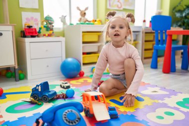 Anaokulunda yerde oturan beyaz bir kız telefon oyuncağıyla oynuyor.