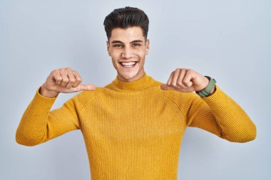 Young hispanic man standing over blue background looking confident with smile on face, pointing oneself with fingers proud and happy. 