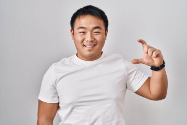 Young chinese man standing over white background smiling and confident gesturing with hand doing small size sign with fingers looking and the camera. measure concept. 