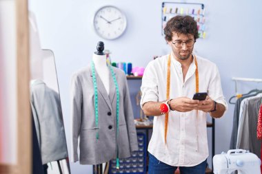 Young hispanic man tailor using smartphone at clothing factory