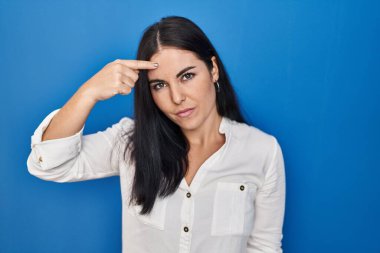Young hispanic woman standing over blue background pointing unhappy to pimple on forehead, ugly infection of blackhead. acne and skin problem 