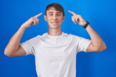 Caucasian blond man standing over blue background smiling pointing to head with both hands finger, great idea or thought, good memory 