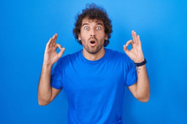Hispanic young man standing over blue background looking surprised and shocked doing ok approval symbol with fingers. crazy expression 