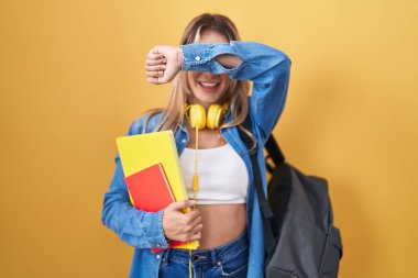 Young blonde woman wearing student backpack and holding books smiling cheerful playing peek a boo with hands showing face. surprised and exited 