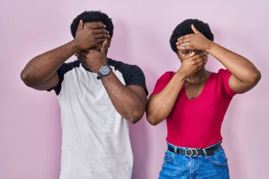 Genç Afro-Amerikan çifti pembe arka planda dikiliyor gözlerini ve ağzını elleriyle kapatıyor, şaşırmış ve şok olmuş. duygu gizleme 
