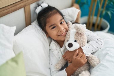Adorable hispanic girl hugging teddy bear lying on bed at bedroom